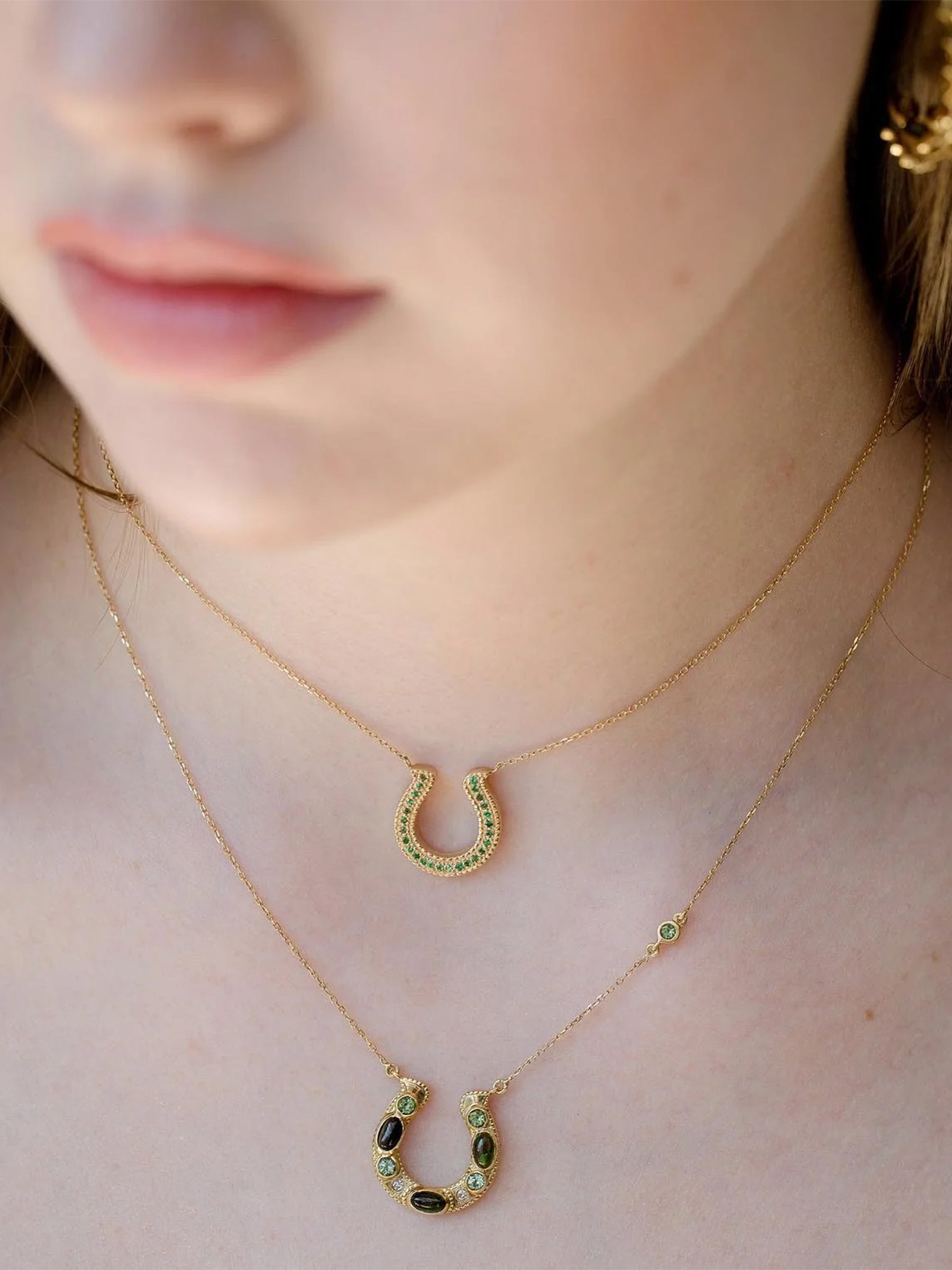18k gold green tsavorite horseshoe necklace worn on model, minimal fine jewelry layered with another horseshoe necklace a close up view.