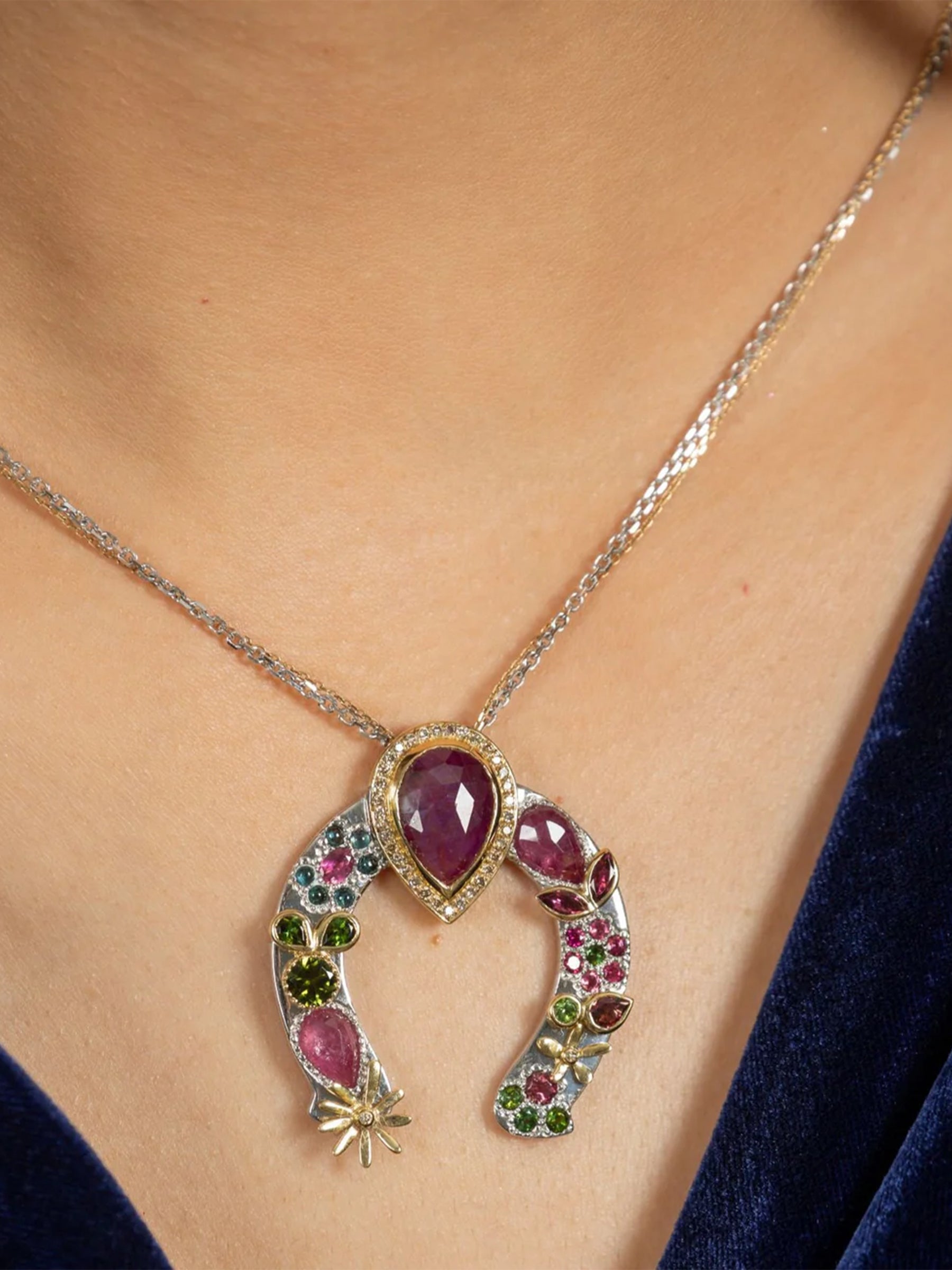 on-model close up view of horseshoe necklace with red sapphire center, tourmaline and sapphire clusters, pavé diamonds, and gold leaf detailing.