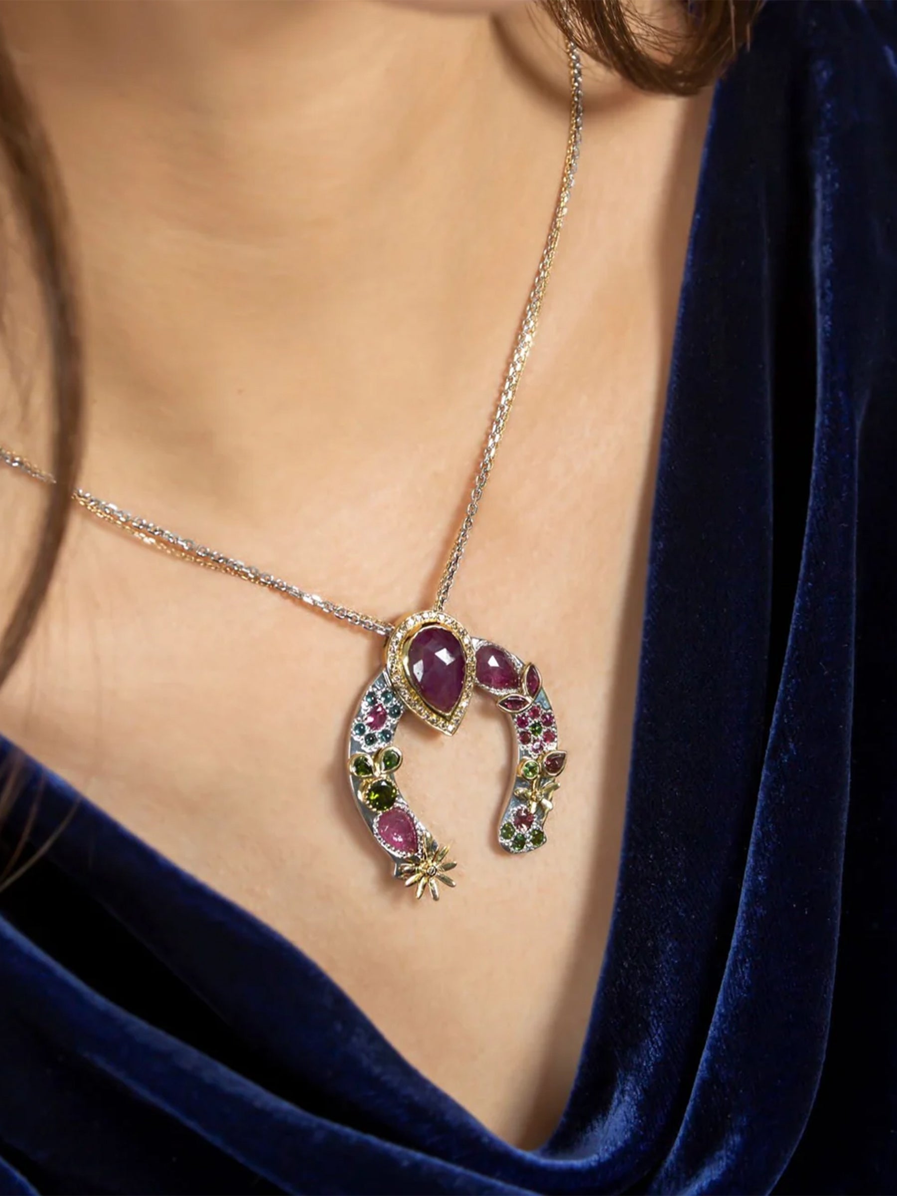 on-model angle view of horseshoe necklace with red sapphire center, tourmaline and sapphire clusters, pavé diamonds, and gold leaf detailing.